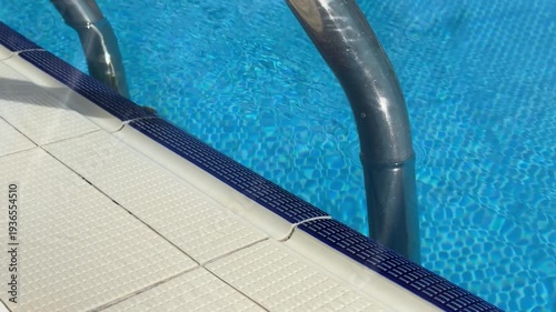 Aluminum ladder descends elegantly into crystal-clear hotel pool water. Sleek metallic steps gleam under sun, inviting safe entry for swimmers. Modern poolside design.