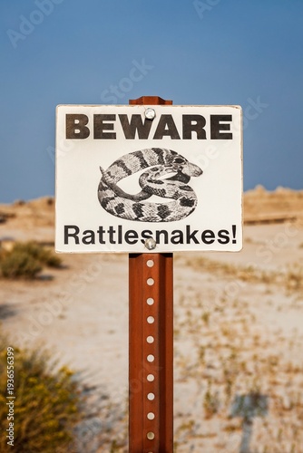 Close view of a Beware Rattlesnakes sign against blue sky and dry prairie in Badlands National Park, South Dakota, highlighting safety messaging and wildlife hazard awareness.