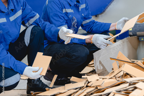 Caucasian male technician and Black female factory worker inspecting cardboard packaging scrap from production line, demonstrating industrial recycling process and environmental waste management.