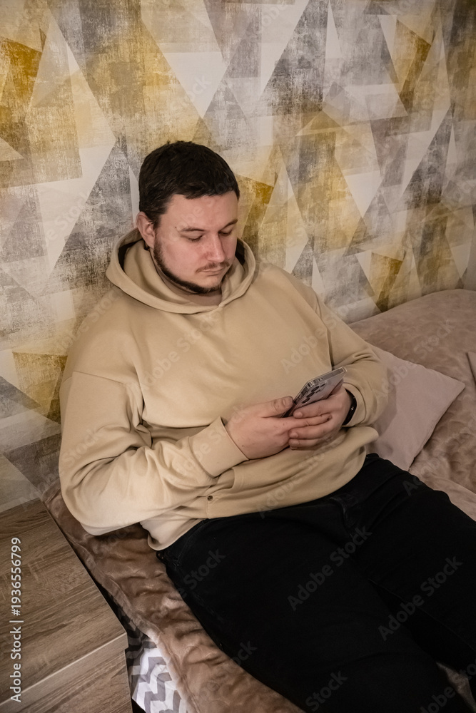 Obraz premium A man wearing a beige hoodie and black pants relaxing on a couch while using a mobile device