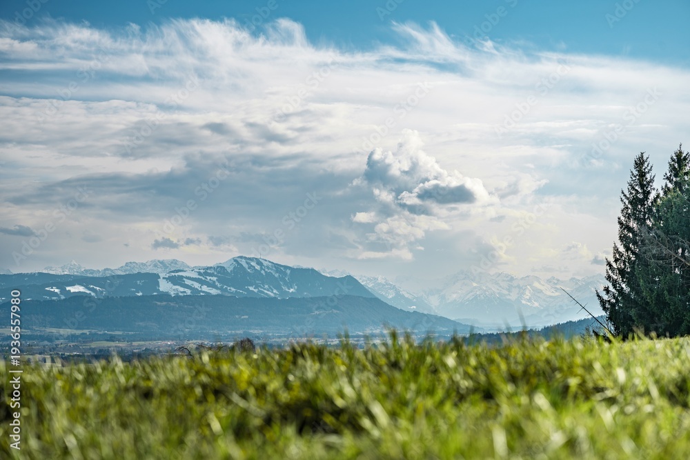 Obraz premium View from Mariaberg above Kempten toward the Allgäu Alps under mixed clouds, green meadow in the foreground and partially snow-covered alpine peaks in the distance.