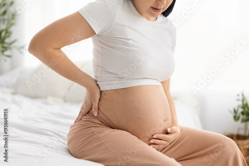 Foto Close up of pregnant woman holding belly and touching lower back while sitting on bed at home