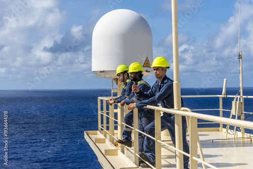 Ship crew members resting above the navigation bridge beside a satellite antenna dome, looking toward the ocean horizon.