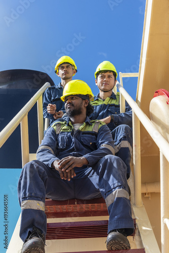 Three seafarers in protective clothing and safety helmets taking a short break while sitting on metal stairs outside the superstructure of a cargo ship.