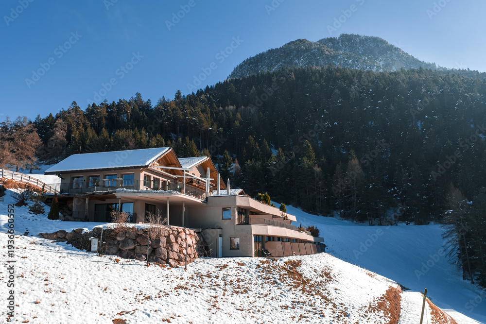 Fototapeta premium A boutique alpine hotel in South Tyrol Dolomites sits on a snowy hillside, with chalet roofs, stone terraces, conifer forests, and a sunlit peak in crisp morning light.