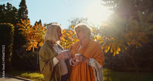 Two senior female friends talking while walking in autumn park with coffee
