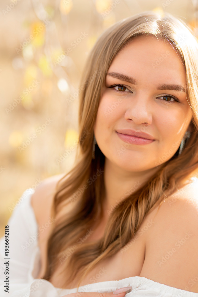 Naklejka premium Close-up portrait of a young woman with flowing brown hair and a gentle expression, bathed in warm, diffused sunlight filtering through foliage