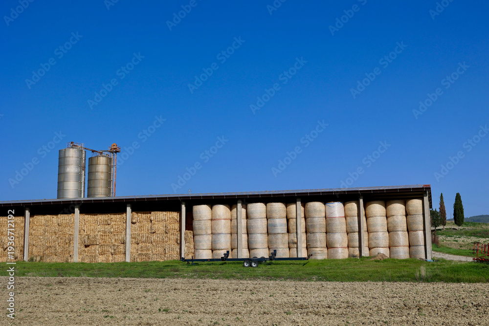 Fototapeta premium Hay Bales Stored in Agricultural Barn with Silos in Tuscan Countryside, Italy