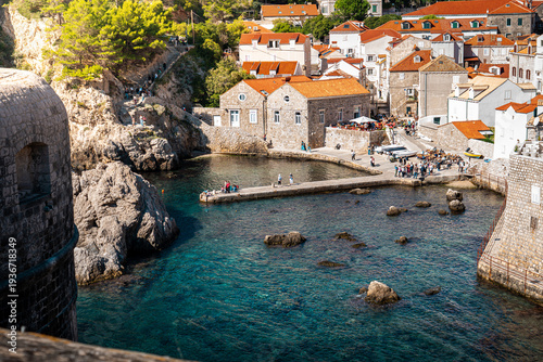landscape shot of harbour beach on Adriatic Sea in dubrovnik old town croatia with red roof houses and clear blue water with tourists walking around and bright green trees in summer with sunlight 