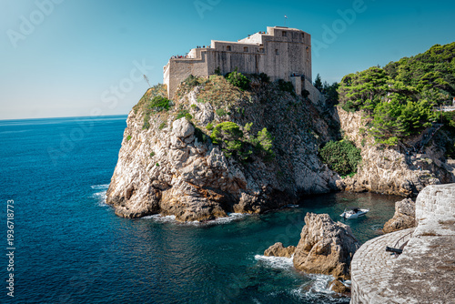 landscape shot looking up at fort Lovrijenac in dubrovnik Croatian coast on the Adriatic Sea with trees and clear blue clouded skies and rocks in the sea in summer
