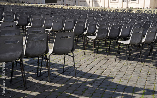 Rows of black plastic chairs arranged on cobblestone pavement in an outdoor setting with shadows cast by sunlight