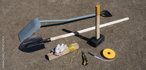 Professional construction hand tools including shovels sledgehammer measuring tape and gloves lying on dry sandy ground