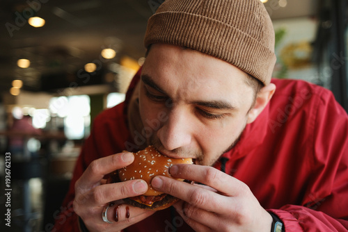 Close up of a young man biting a juicy beef burger with bacon on a sesame bun while sitting in a fast food restaurant. Concept of casual dining, street food and quick urban meal
