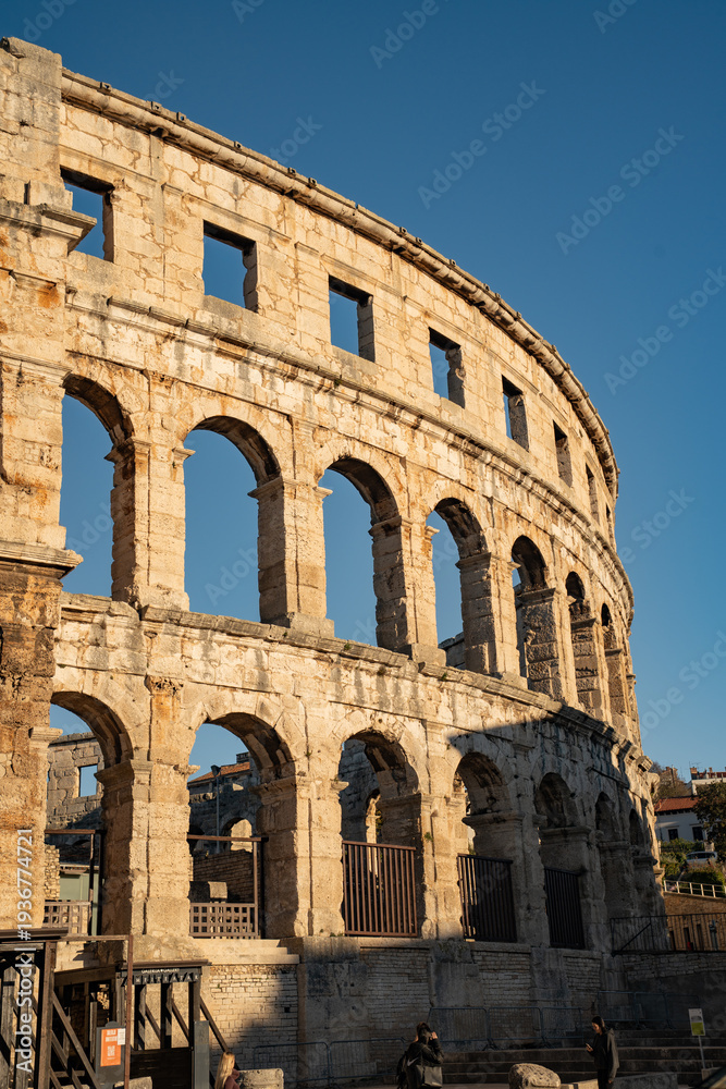 Fototapeta premium Epic Pula arena ancient Roman architecture vertical panorama. Cinematic low angle view of historic monument amphitheater ruins, limestone columns, arcade arches and golden sunset sky. 