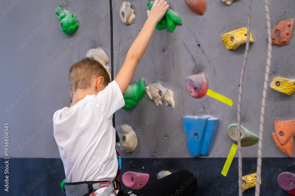 Fototapeta premium Young Boy Climbing an Indoor Bouldering Wall with Colorful Holds at Sport Gym Wearing a Harness