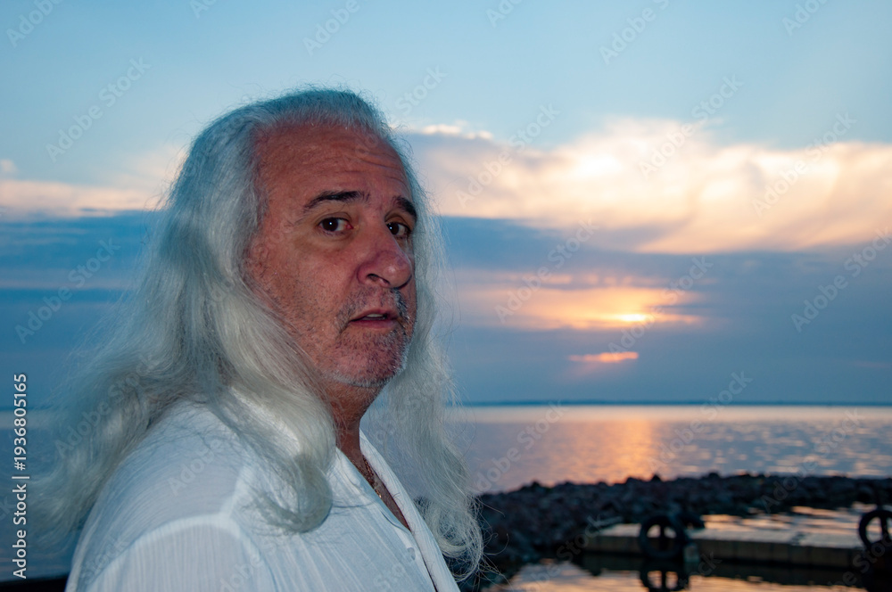 Fototapeta premium Senior lifestyle. Natural aging beauty. Mature male grooming. Mature man by ocean horizon during sunset. Healthy long hair at older age. Authentic masculine style. Retired man enjoying evening