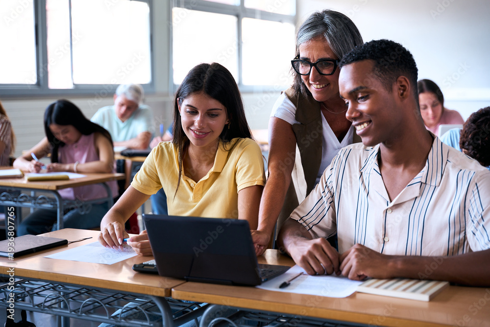 Fototapeta premium A mature female educator assists a young man and woman during a computer lesson. They are smiling and engaged while looking at the screen in a bright school environment.