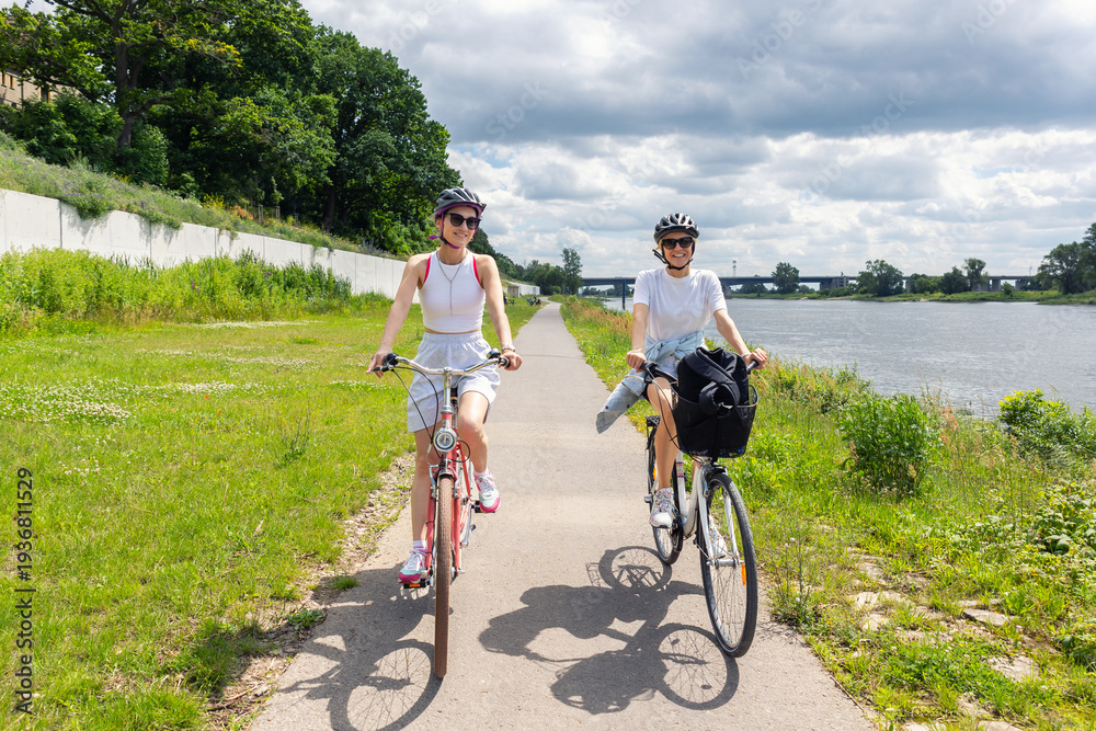 Fototapeta premium Women riding bicycles along riverside path during summer day with green park landscape and cloudy sky. Active lifestyle, friendship, outdoor recreation healthy activity travel freedom leisure cycling