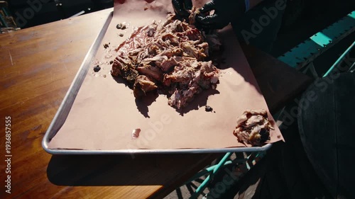 Black-gloved hands gather and consolidate shredded pulled pork on pink butcher paper in a metal tray in an overhead slow-motion shot