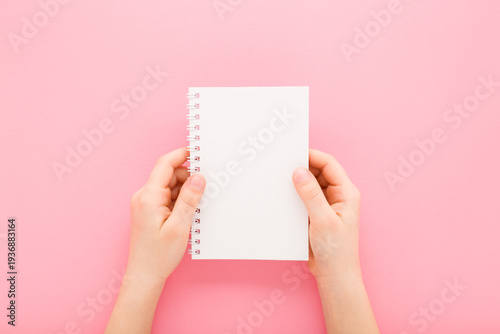 Little child girl hands holding white copybook on light pink table background. Pastel color. Closeup. Empty place for text. Point of view shot. Top down view.
