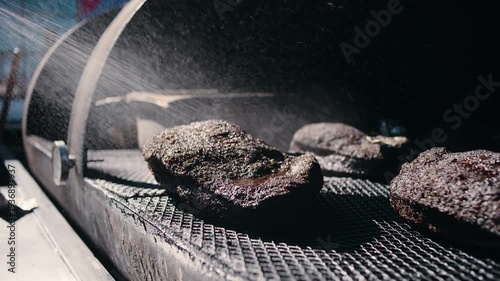 A pitmaster in a canvas apron walks past a large outdoor woodpile carrying a split log of firewood for the smoker in daylight