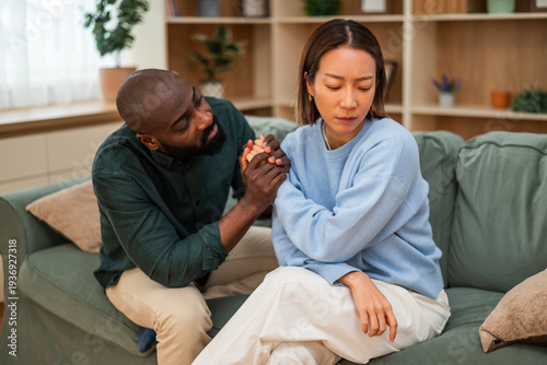 Multiracial couple sitting on sofa at home and arguing. Woman is angry and man is apologizing to her. Relationship difficulties.