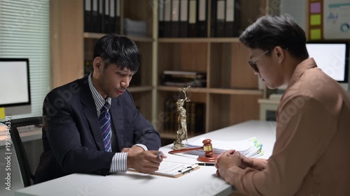 Two men are sitting at a desk, one of them writing on a piece of paper. Scene is serious and professional, as the men are likely discussing important matters