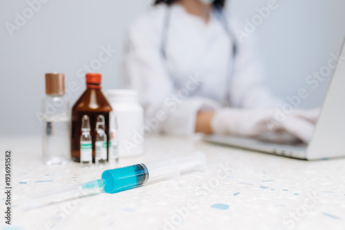 Medical syringe with blue vaccine liquid on clinic desk and doctor working on laptop in background