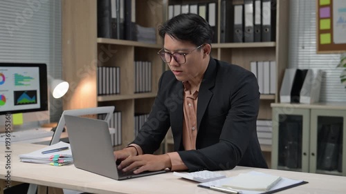 A man in a suit is typing on a laptop in front of a monitor. The room is filled with bookshelves and a potted plant. The man is focused on his work, possibly in a professional setting