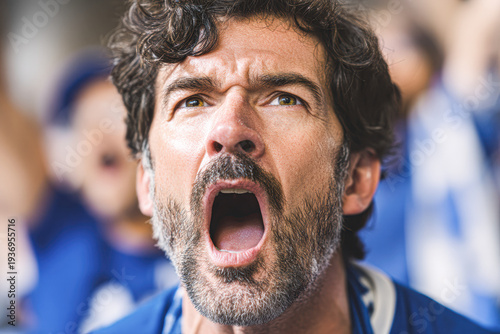A man with curly hair and a beard passionately shouts, possibly cheering or reacting emotionally, with a blurred crowd in the background.