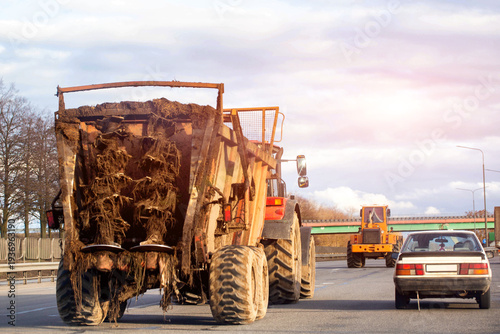 Large agricultural tractor with trailer full of roots and soil travels on multilane highway alongside other vehicles under cloudy sky during daytime. Copy space for text