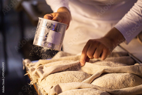 Hands of baker dusting fresh sourdough bread dough with flour using sifter for artisan preparation in professional kitchen setting.