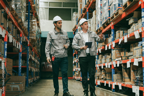 Konstfotografi Two male warehouse workers are talking in the moving aisle area and jointly checking the orderliness of parcels placed on pallets on shelves in a storage area in an industrial and logistics warehouse
