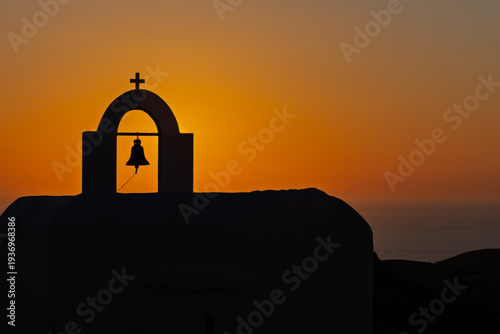 View of a the silhouette of a Greek Orthodox Church, with a bell and a cross at the top and a spectacular orange sunset in the background in Ios cyclades Greece