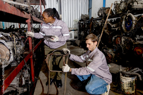 Harmony diversity people, white guy and African woman workers in safety protective uniform working together, use cart to transport machinery engine spare parts. Engineers checking stock at warehouse.