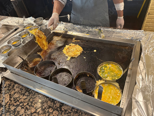 Cook preparing omelet on industrial griddle in restaurant kitchen. Professional chef using spatula to fry egg dish. Culinary process of food preparation in hotel breakfast service.