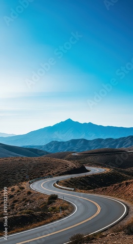 Winding asphalt road stretching across a hilly, undeveloped landscape, leading toward a distant mountain range under a vast blue sky, wanderlust, vanishing point, aspiration