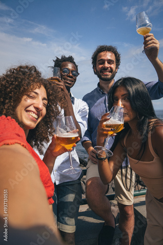 Multicultural friends taking selfie while drinking beer outdoors on sunny seaside day