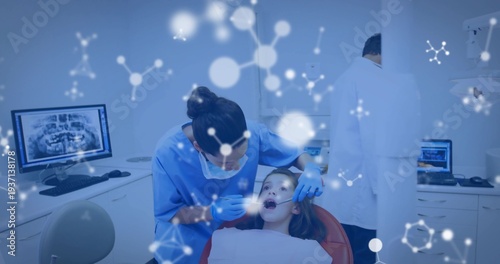 Examining clinician in blue scrubs leaning over child in red dental chair, using mirror and gloves