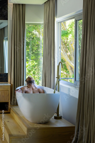 Mature African American man bare-chested reclining in egg-shaped tub in bathroom with gold faucet