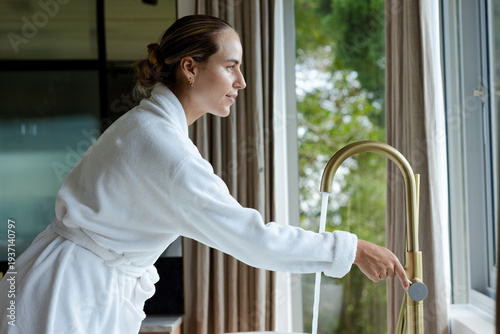 Woman in her 20s wearing white robe at kitchen sink operating brass gooseneck faucet, water flowing