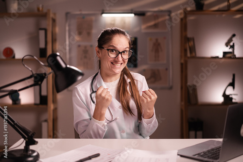 Portrait of female doctor or medical student intern with stethoscope celebrating win victory sitting at office desk looking at camera working during night shift at hospital