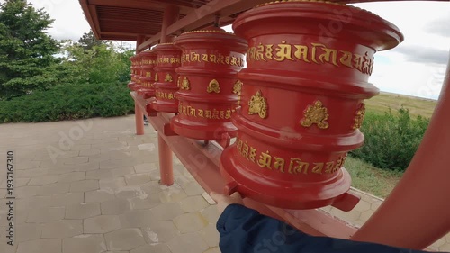 POV a traveler's hand spinning a red Buddhist prayer wheel with gold mantras. Conveys spirituality, travel, and mindfulness in a serene outdoor temple setting with natural light