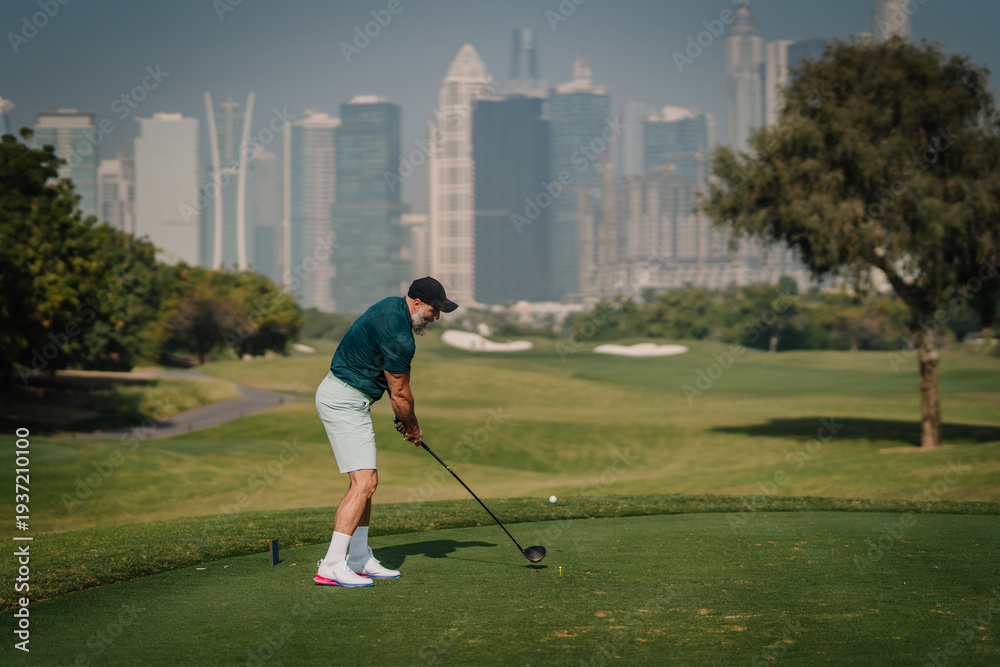 Fototapeta premium Male golfer in green shirt and white shorts prepares to swing golf club on tee box with city skyline and green fairway in the background