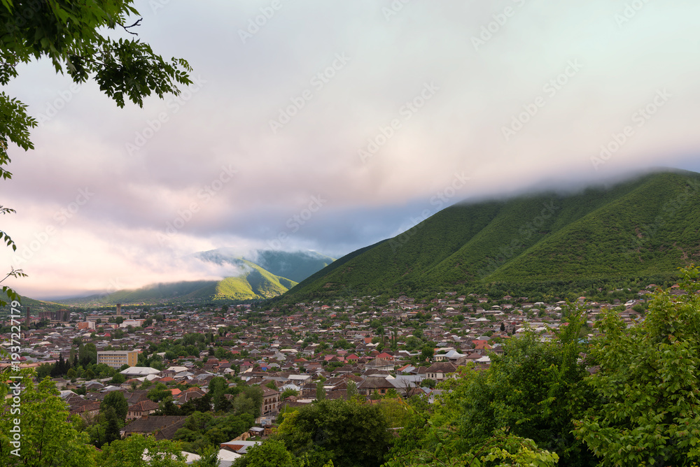 Obraz premium Shaki City, Azerbaijan, nestled in a valley with green mountains partially covered by atmospheric clouds. Overhead view of houses and trees under a soft sky