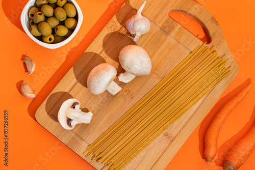 Food ingredients for cooking Italian pasta. Fresh cherry tomatoes, mushrooms, garlic, parsley, olives, spaghetti, olive oil on orange background top view