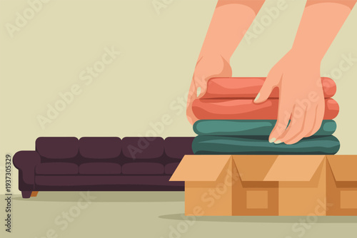 Hands stacking folded clothes in a cardboard box near a couch, home organization concept