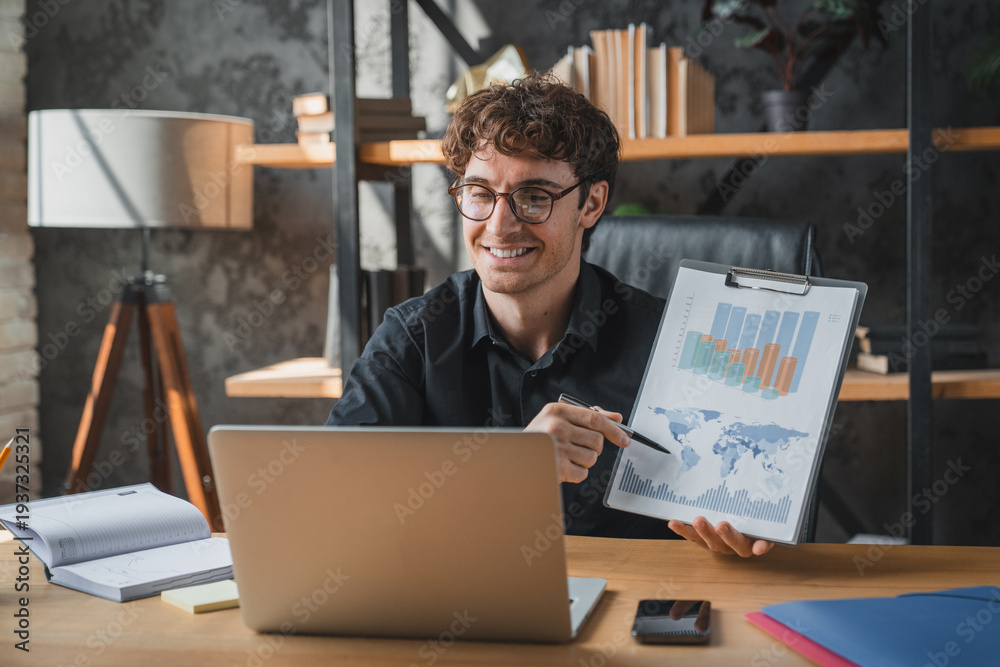 Fototapeta premium Man, video call laptop in office for paperwork, remote work and graphs for report. Freelancer, business documents and tech for project management, finance agency, giving consultation, conference app
