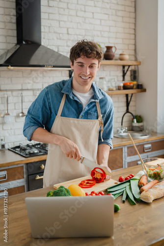 Smiling Caucasian male chef in apron preparing salad in modern kitchen interior with organic vegetables and laptop. Food blog at home, new recipe to cook healthy dinner at home and diet. Vertical shot