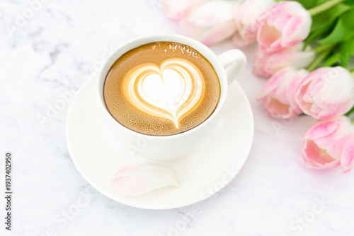 Cup of coffee with latte art heart and pink tulips on white marble background. 